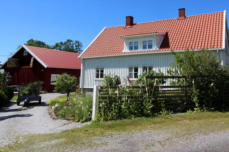 Ett vitt trähus med rött tegeltak omges av en trädgård och ett lågt staket, vilket skapar en scen värdig bilder. I närheten står en röd lada under en klarblå himmel, och en grusad uppfart leder till båda byggnaderna.