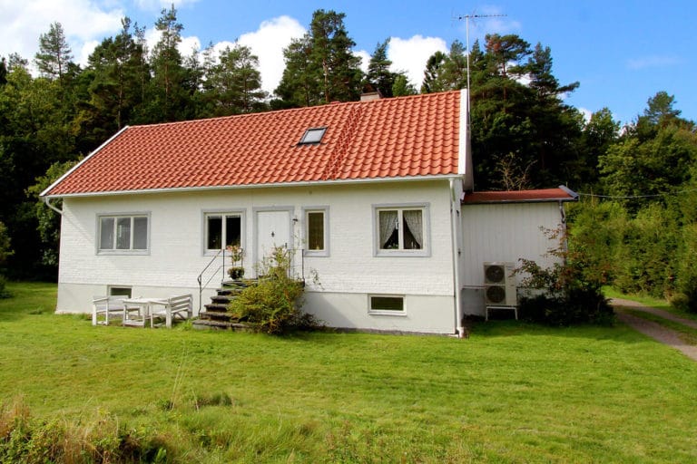 Ett vitt hus med rött tegeltak ligger på en gräsmatta omgiven av träd. En trappa leder till ytterdörren och bilderna visar en liten uteplats med sittplatser och en sidoutbyggnad med två luftkonditioneringsenheter.