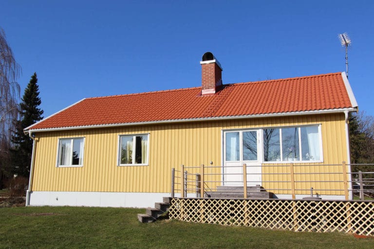 Ett gult envåningshus med rött tegeltak, vitkarmade fönster, skorsten och metallräcke står på en gräsmatta under en klarblå himmel - perfekt för att fånga vackra bilder av ett lugnt liv.