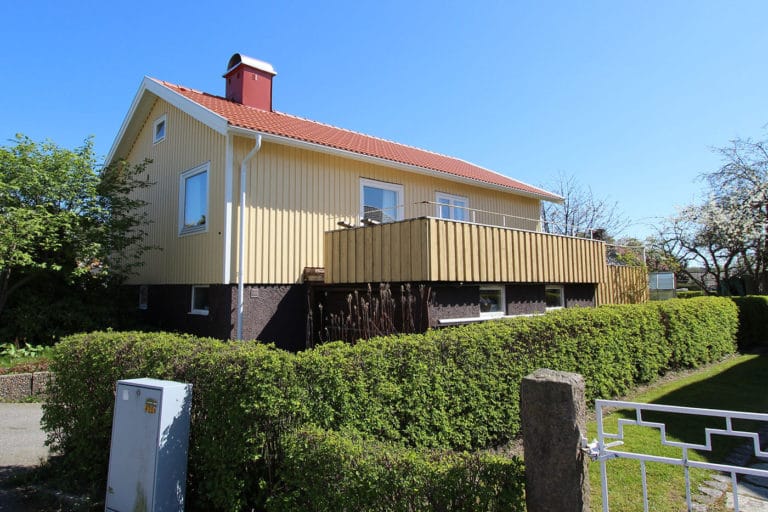 Tvåvåningshus med gul fasadbeklädnad, rött tegeltak och stor balkong. Fastigheten, som finns med på flera bilder, avgränsas av en grön häck och träd, och i förgrunden syns en metallgrind och en elbox.