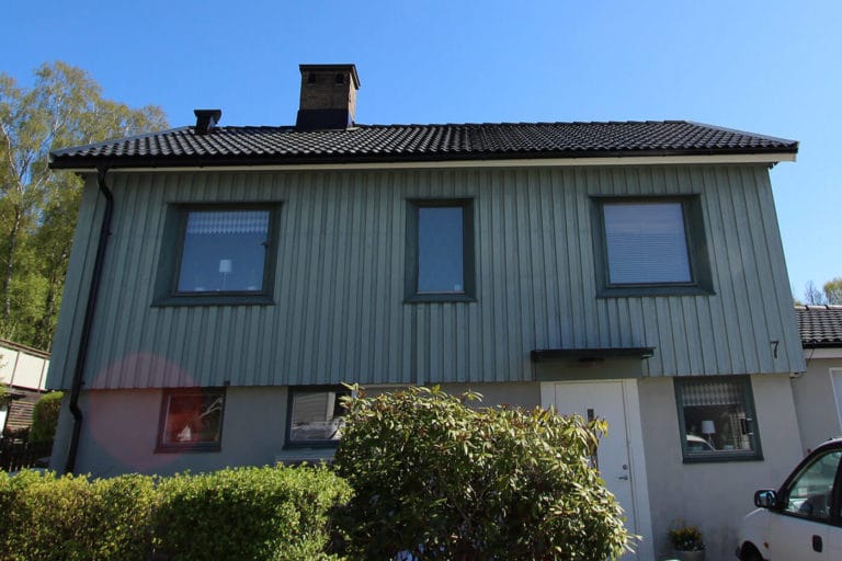 Ett tvåvåningshus med vertikal grön träfasad och ett mörkt tegeltak, med fyra fönster på övervåningen och en vit entrédörr på undervåningen, omges av gröna buskar och buskage - perfekt för att inspirera till schöne bilder.