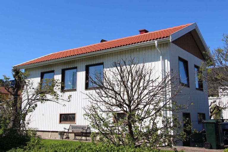 Ett vitt tvåvåningshus med rött tegeltak och flera stora fönster, omgivet av träd och buskar en solig dag - perfekt för att ta bilder. En bänk står nära ytterväggen.