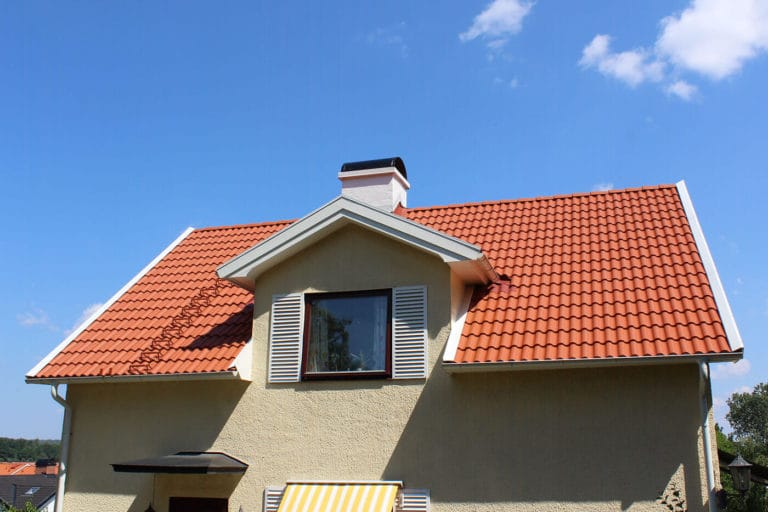 Ett beige hus med rött tegeltak, skorsten, vita fönsterluckor och en gulrandig markis står under en klarblå himmel med några moln - en scen som är perfekt för bilder eller heminspiration.