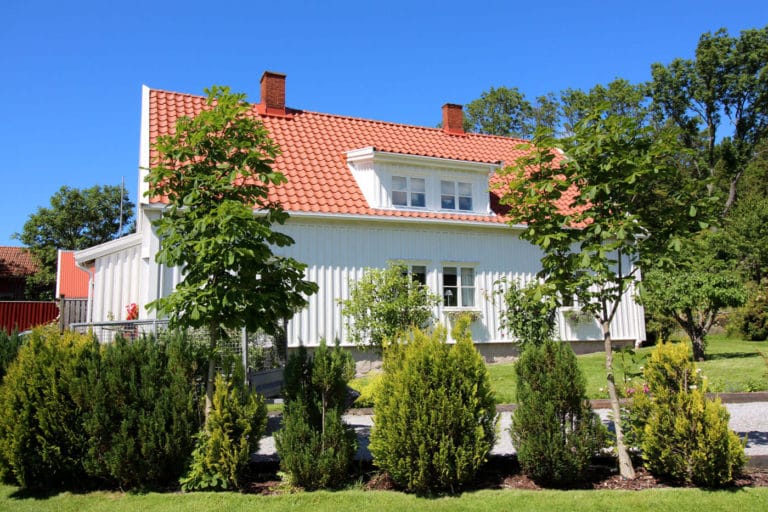 Ett vitt hus med rött tegeltak står bakom en rad unga träd och buskar en solig dag, omgivet av grönt gräs och blå himmel - perfekt för att ta vackra bilder av fastigheten.
