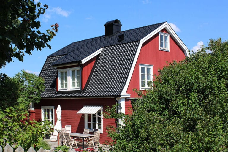 Ett rött trähus med vita lister och svart tegeltak omges av gröna träd. På uteplatsen, som finns med på många bilder, står ett bord, stolar och ett vitt parasoll under en klarblå himmel.