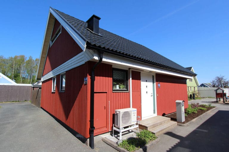 Ett rött trähus med svart tegeltak, vita lister och en liten veranda på framsidan. En luftkonditioneringsenhet är monterad utanför nära dörren. Den klarblå himlen ger vackra bilder av denna charmiga scen.
