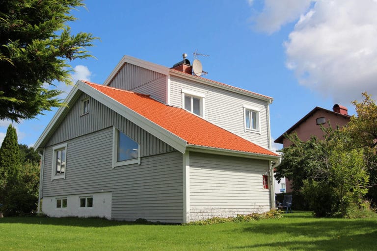Ett vitt tvåvåningshus med rött sluttande tak ligger på en grön gräsmatta under en delvis molnig himmel. En parabolantenn för TV är monterad på taket och grannhusen syns i bakgrunden på dessa bilder.