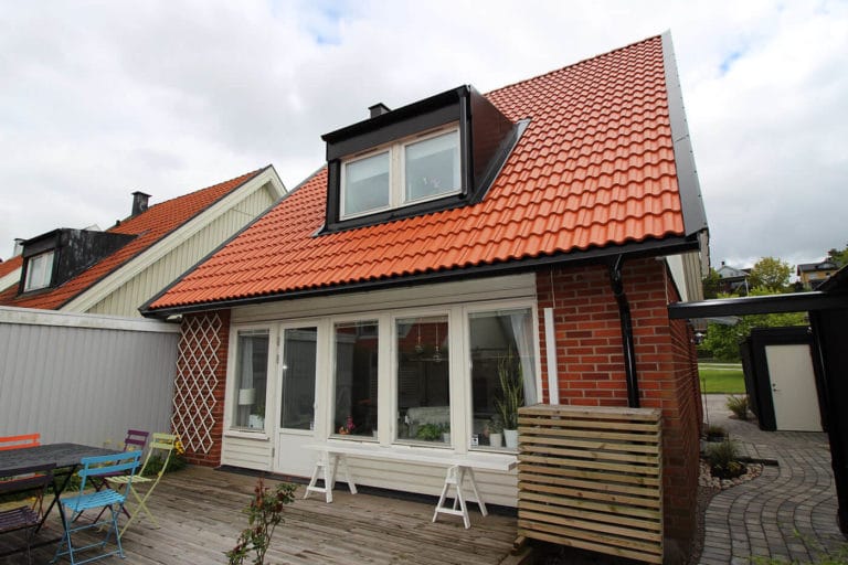 Ett litet hus med rött tegeltak och takkupa, fönster och dörrar med vita ramar, rött tegel och vit fasadbeklädnad samt en uteplats i trä med utemöbler - perfekt för att fånga charmiga bilder under den molniga himlen.