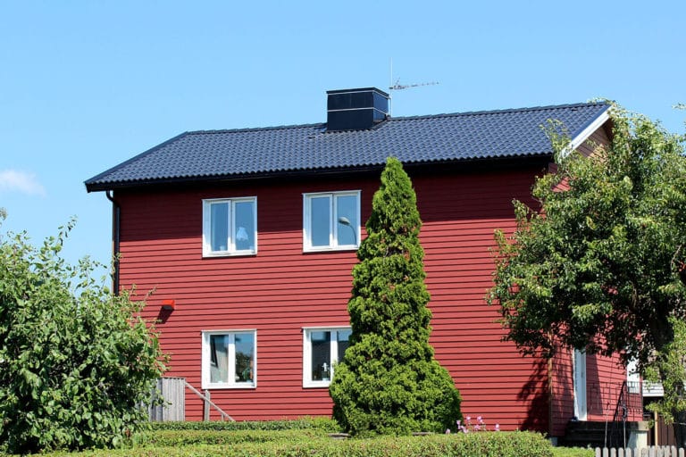 Ett rött trähus i två våningar med vit dekor och mörkblått tegeltak, omgivet av gröna träd och buskar under en klarblå himmel, perfekt för bilder som fångar fridfulla landsbygdsscener.