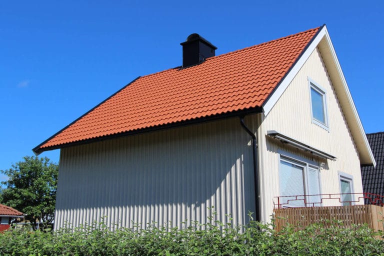 Ett beige hus med rött tegeltak och svart skorsten, som förekommer i många bilder, har två stora fönster och en liten balkong och står bakom en grön häck under en klarblå himmel.