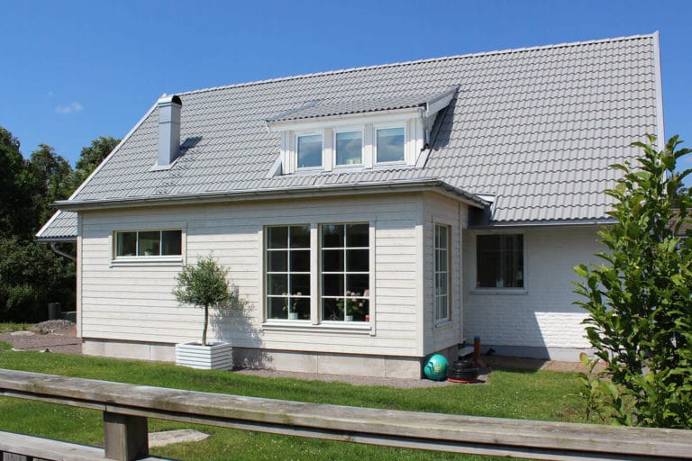 Ett ljust hus med grått tegeltak, stora fönster och en liten takkupa ligger på en gräsmatta med ett träd och en trädgårdsslang - perfekt för att fånga vackra bilder under den klarblå himlen.