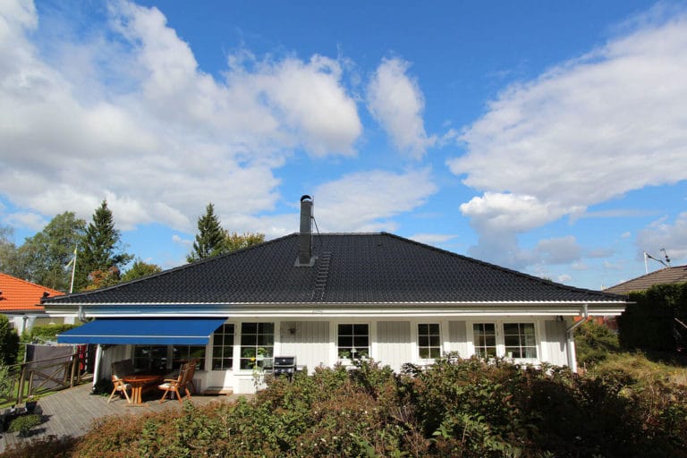 Ett vitt envåningshus med mörkt valmat tak och en blå infällbar markis täcker en uteplats med utemöbler. Gröna buskar kantar förgrunden under den delvis molniga himlen - perfekt för att fånga fantastiska bilder.