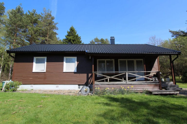 Ett envånings trähus med mörkt tak, vitkarmade fönster och en liten täckt veranda med trappsteg, som ligger på en gräsmatta med träd i bakgrunden en klar dag - perfekt för att fånga vackra bilder.